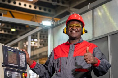 Cheerful African American CNC Machine Operator Standing Next To CNC Controller Console And Giving Thumb Up. Portrait Of Black Machine Operator In Red Helmet, Safety Goggles, Hearing Protectors And Work Uniform.
