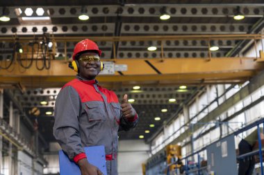 Cheerful African American Blue-Collar Worker Giving Thumbs Up In A Factory. Portrait Of Black Worker In Red Helmet, Safety Goggles, Hearing Protectors And Work Uniform. 