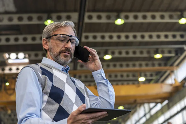 Chief Executive Officer Talking On Smartphone And Looking At Digital Tablet Screen In Industrial Interior. Waist Up Portrait Of Confident, Elegant, Gray-Haired CEO Or Businessman. Man Standing And Using Smartphone And Digital Tablet.