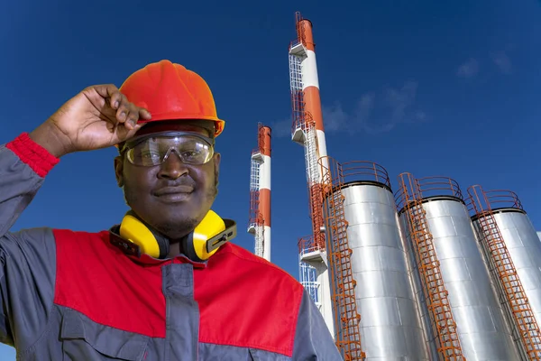 Black Worker In Personal Protective Equipment Standing In Front Of District Heating Plant. Young African-American Power Plant Or Oil Worker In Hardhat Standing In Front Of Industrial Storage Tanks.