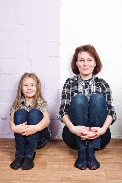 Mother with the daughter in jeans and a shirt sit on a floor against a wall from two flowers — Stock Photo, Image