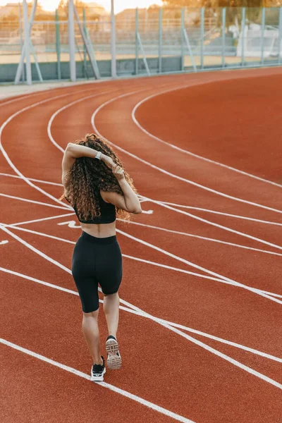 Siyah spor giysili, permalı saçlı bir kadın stad üzerinde geriliyor.