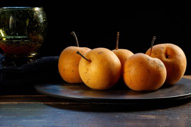 Çin Nashi armutları, siyah tabakta yuvarlak kahverengi meyveler ve siyah arka planda klasik şarap kadehi. Alçak tuş.