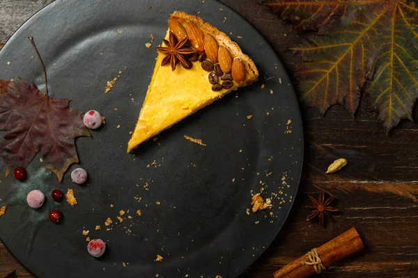 The last piece of pumpkin cheesecake on a black stone plate. The concept of a festive table. Top view on rustic wooden background with autumn leaves and spices.