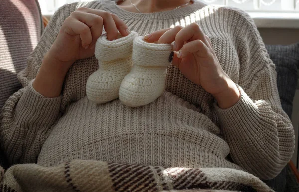 Mujeres embarazadas en un suéter beige sostiene botines blancos de punto bebé en su barriga — Foto de Stock