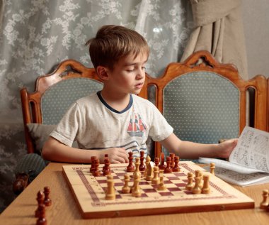 Sarışın bir çocuk bir satranç kitabı okuyor. Önünde taş olan bir satranç tahtası. Seçici odak.