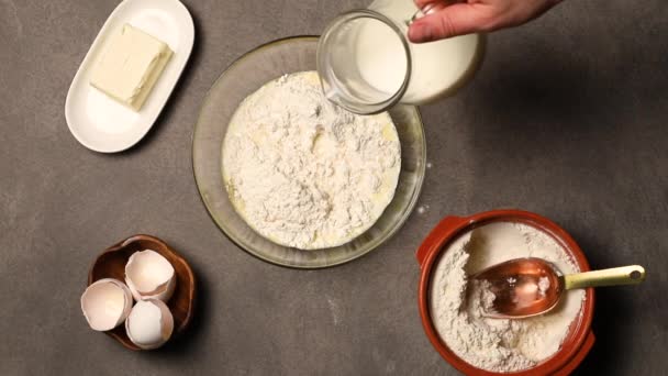 Förberedelse av smet för pannkakor. Kocken häller mjölk ur en kanna med en skål slagna ägg och mjöl. Ovanifrån. — Stockvideo