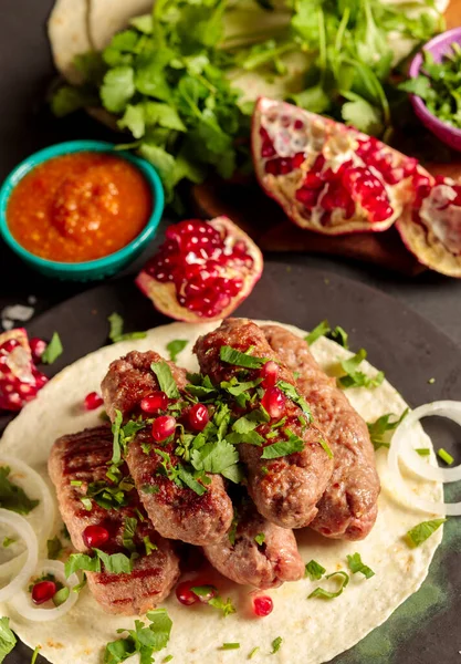 Meat kebabs on tortillas with chopped herbs, pomegranate seeds and hot sauce. Close-up, on a gray background, vertical