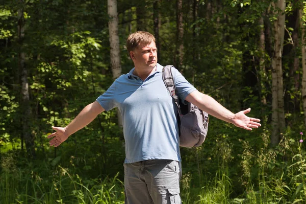 A hitchhiker tourist with a backpack on his shoulder throws up his hands in surprise on an empty road against the background of a forest.