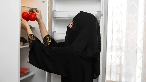 穿着民族服装的穆斯林妇女在新的空冰箱里放红色多汁的西红柿 — 图库照片
