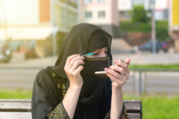 街の公園のベンチで民族衣装を着たイスラム教徒の女性が鏡を見て彼女のまつ毛を描きます — ストック写真