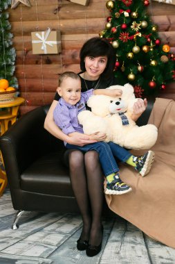 Annem küçük oğlunu kollarında tutuyor. Deri bir koltukta oturuyor. Bir Noel ağacının arka planında..