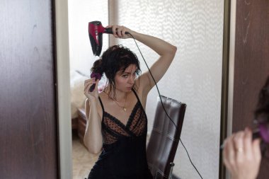 Aynada ıslak saçlı yetişkin bir esmer saçını saç kurutma makinesiyle kurutur..