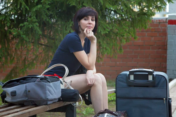A happy brunette woman is sitting bench at the train station with a lot of bags and a suitcase waiting for her train.