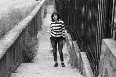 Çizgili kazaklı bir kadın metal çit boyunca kaldırımda yürüyor..