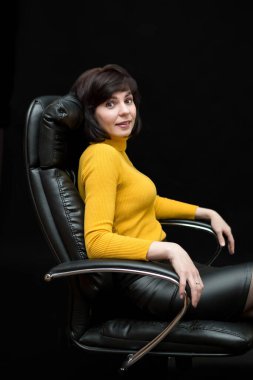 35-40 yaşlarında esmer bir kadın büyük bir deri koltukta siyah arka planda oturuyor..