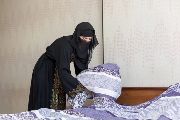 Muslim immigrant woman making bed in bedroom.,
