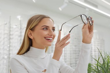 Optik Dükkan. Gözlük arayan, vitrinin yanındaki kadın. Gülümseyen Yüzlü Mutlu Kız Optik Mağazasında Yeni Gözlük Seçiyor. Arka planda Farklı Gözlük Modelleri Gösterimi