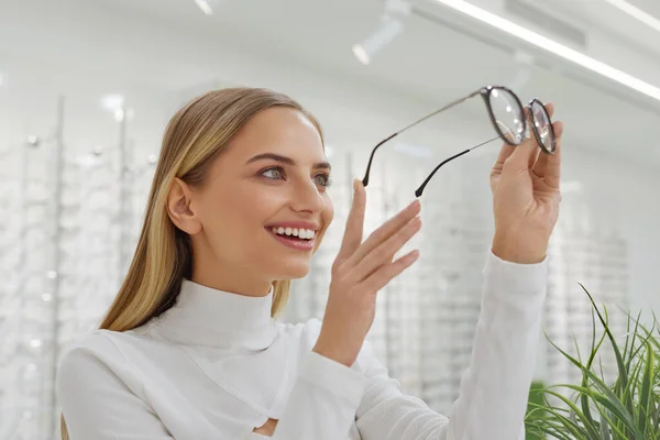 光学店 眼鏡を探しているショーケースの近くの女性 Optics Storeで新しいメガネを選ぶ笑顔の幸せな女の子 背景に異なるモデルのメガネを持つショーケース — ストック写真