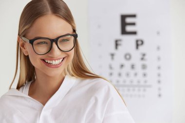 Optometri ve Vizyon. Ofisteki gülümseyen kız. Sağlıklı, gülen, güzel yüzlü bir kadın. Görsel Göz Testi Çizelgesinin Önündeki Genç Kadının Portresi. Görsel Sağlık Konsepti