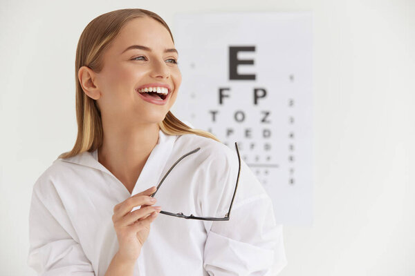 Optometry And Vision. Smiling Girl At Ophthalmologist Office. Portrait Of Beautiful Woman Laughing Out Loud and Holding Glasses with Visual Eye Test Chart On Background. High Resolution Image