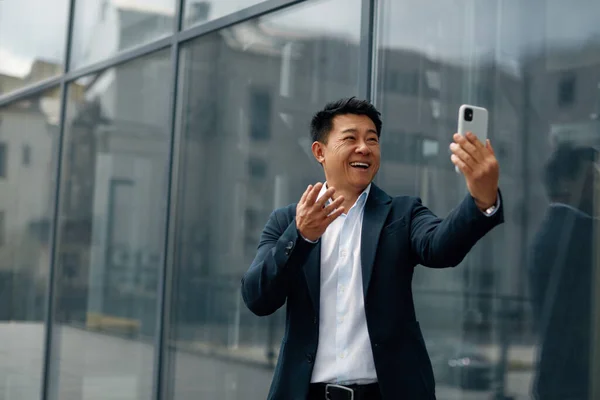 Asian Man Gesturing, Chatting Video Call Using Smartphone on City Street. Confident Smiling Male Worker Using Mobile Phone Alone. Corporate Lifestyle Concept
