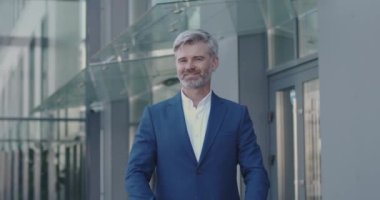 Kameraya Tek Başına Bakarken Gülümseyen ve Kollarını çaprazlayan Yetişkin İş Adamının Portresi. Modern Cam Ofis Merkezi 'nin yakınında başarılı bir erkek işadamı duruyor. İş İnsanları Konsepti 