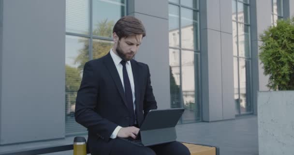 ラップトップの屋外でベンチで働く深刻なひげそりビジネスマン 男性シニアオフィスワーカーが現代の都市背景の職場で街に座る — ストック動画
