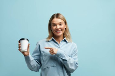 Kahveyi işaret eden gülümseyen kadın izole edilmiş. Sarışın, elinde sıcak kahve tutan bir kadın, tek kullanımlık içecek bardağını işaret ediyor. Kapalı Stüdyo Çekimi Mavi Arkaplanda İzole Edildi 