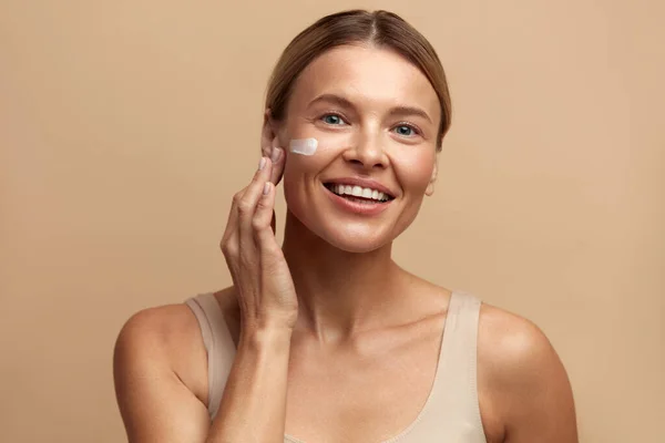 Skin Care Woman Applying Cream on Cheek. Closeup Of Beautiful Smiling Girl Putting Cream On Fresh Soft Pure Skin. Portrait Of Woman With Natural Makeup Applying Beauty Cosmetics Product