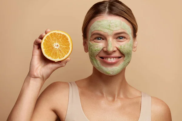 Laughing Face Mask Woman Hold Orange. Positive Woman with Clay Face Mask. Happy Young Woman Looking at Camera with Slice of Citrus Fruit. Concept of Skincare. Isolated on Beige Background
