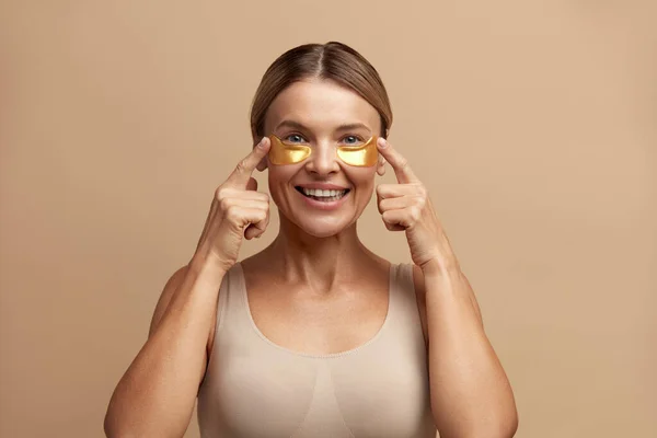 Woman Beauty Face With Mask Under Eyes. Closeup Of Beautiful Smiling Girl With Gold Patches, Golden Collagen Mask On Facial Skin. High Resolution