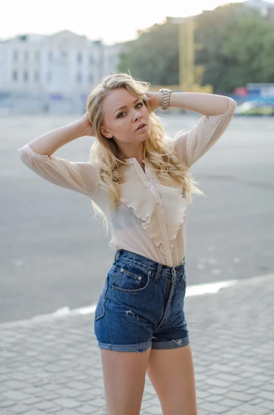 Chica rubia en pantalones vaqueros azules y blusa blanca posando en la calle Fotos de stock libres de derechos