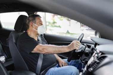 Senior in his 70s driving a car wearing face mask for protection against corona virus