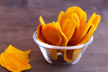 Bir kasede cips. Ahşap arka planda cam kasede kurutulmuş mango ve fotokopi uzayı. Süper yemek, vejetaryen, vejetaryen gıda konsepti. Portakal kurutulmuş mango dilimleri. Sağlıklı çerez.