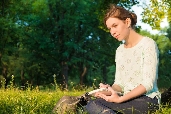 年轻漂亮的女孩坐在一个公园里，在笔记本上书写 — 图库照片