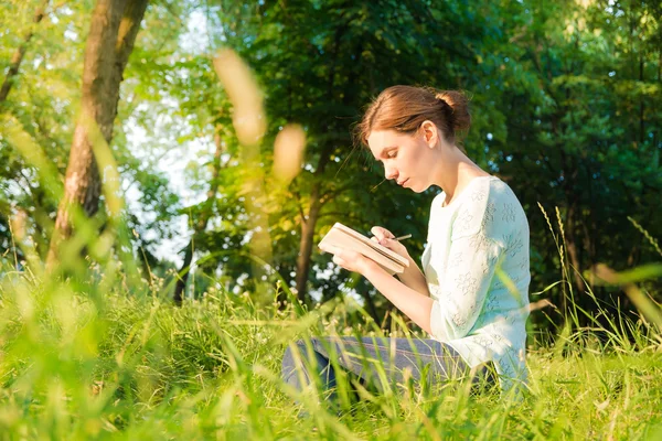 Ung vacker flicka sitter i en park och skriva i en anteckningsbok — Stockfoto