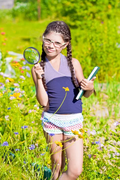 Girl with magnifying glass and book stays among flowers — Stock Photo, Image