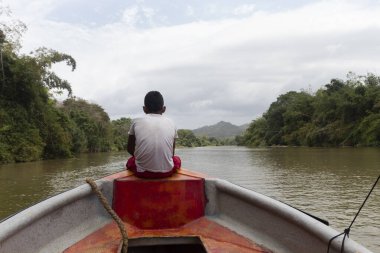 Yerli bir çocuk turist teknesinin önünde oturmuş arka planda bulutlu Sierra Nevada dağları olan Palomino Nehri manzarasını seyrediyor.