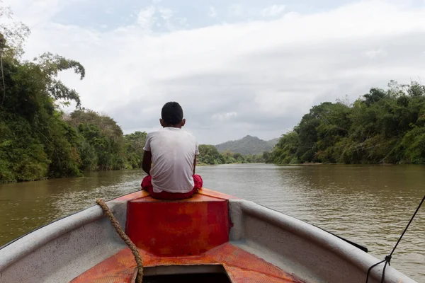 Yerli bir çocuk turist teknesinin önünde oturmuş arka planda bulutlu Sierra Nevada dağları olan Palomino Nehri manzarasını seyrediyor.