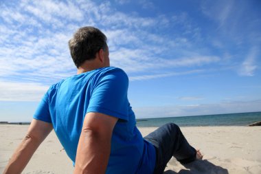 Kumsalda kumsalda oturan mavi gömlekli adam.