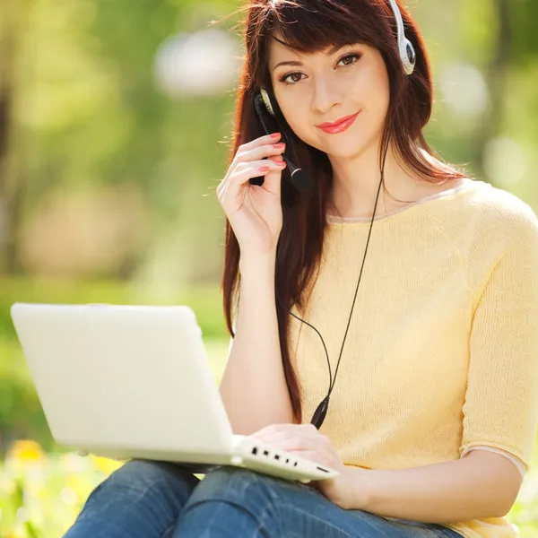 Carino donna in auricolare con computer portatile bianco nel parco — Foto Stock