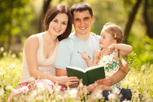 Mãe feliz, pai e filha lêem um livro no parque — Fotografia de Stock