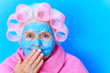 Yüzünde mavi krem nemlendirici maske olan kadın, beyaz saç bigudileri takıyor ve stüdyo arka planında pembe banyo bornozu giyiyor.