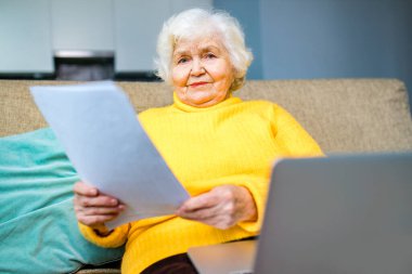 Gümüş saçlı, olgun, mutsuz Rus kadın modern evdeki oturma odasında bankacılık kağıt faturasını tutuyor.