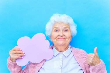 Kar beyazı beyaz saçlı, pembe hırkalı ve mavi gömlekli olgun bir kadın stüdyo arka planında kağıt bulutu tutuyor.