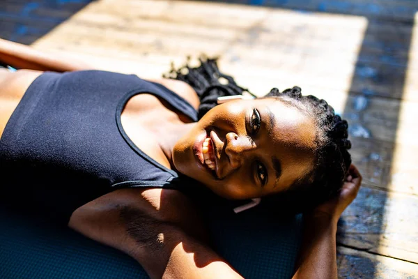 Authentische afrikanisch-amerikanische Frau in schwarzem, sportlichem BH legt sich auf den Boden einer Yogamatte, hört Musik und blickt in die Kamera — Stockfoto