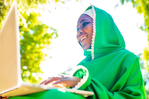 Hispanic kvinna i grön muslim hijab med ljusa make up och näsa piercing arbetar med laptop sitta på gräs utomhus sommarpark — Stockfoto