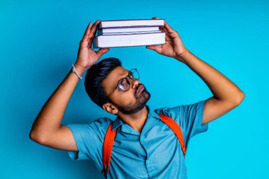 Hintli öğrenci kitaplara bakarken mutlu hissediyor mavi stüdyoda gözlük ve turuncu sırt çantası takıyor.
