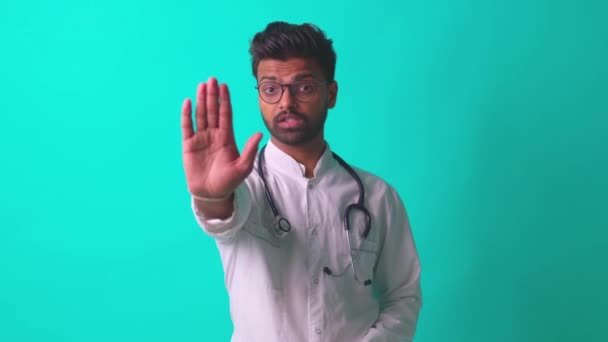 Молодой латиноамериканец-медик носит очки со стетоскопом и рассказывает плохие новости на фоне синей стены студии — стоковое видео